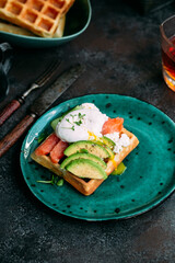 Belgian waffles with salmon, poached egg, avocado and cream cheese on a turquoise plate. Healthy breakfast at the table