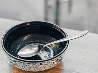 Empty dirty plate after meal with metal spoon inside stands on wooden table near window. Black and white soup plate