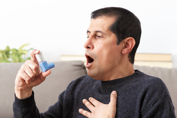 Man holding inhaler with asthma attack suffocating sitting on a couch in the living room at home