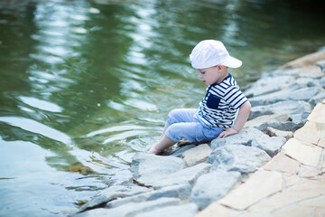Baby in white cap sitting by the pond touches cold water with his leg and admires