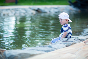 little naughty boy is happy and smiling, sitting on the edge of a pond in a city park, wearing a cap turned back