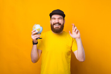 Cheerful young bearded man holding globe and crossing fingers over yellow background
