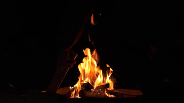 Man puts oak firewood. Puts the log in the fire. Smoldering wooden logs in warm flame. Close-up of sparks from conflagrant bright fire. Orange twisting tongues of fire.Smoldering night bonfire camping