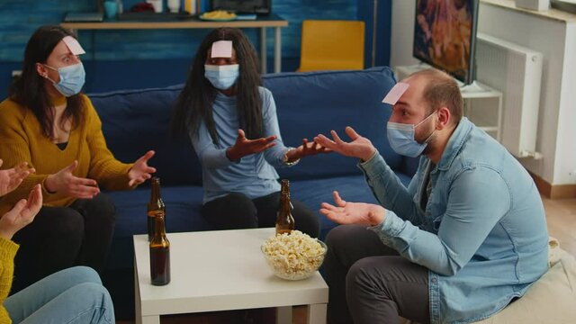 Thoughtful multiethnic friends with sticky notes on foreheads playing name game miming gesturing wearing face mask keeping social distancing preventing covid19 spread. People enjoying bottles of beer