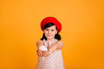 Adorable kid in stylish beret celebrating birthday. Caucasian female child holding cake with candle isolated on yellow background.
