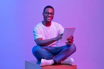 Black Student Guy With Digital Tablet Sitting On Cube Under Neon Light - obrazy, fototapety, plakaty