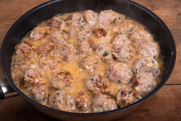 Chicken meatballs with cheese in a creamy sauce in a frying pan on a wooden kitchen table, top view, close-up - Swedish cuisine