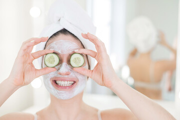 Cheerful woman with a towel on her hair and in a clay face mask and cucumbers on her eyes. Taking care of beauty at home