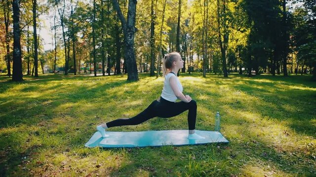 Focused purposeful girl sitting on the floor, blue mat, stretching her arms, bending over, want to be strong, physically healthy, doing yoga in a summer park.