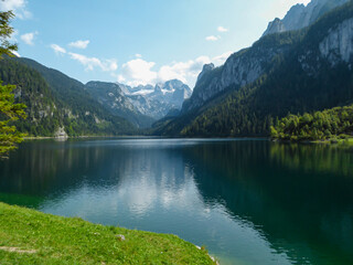 Naklejka na ściany i meble Panoramic view on Gosau lake, with Dachstein glacier in the back in Austrian Alps. The lake is surrounded by high mountains, overgrown with tall trees. Sun reflects on the surface. Serenity and calm