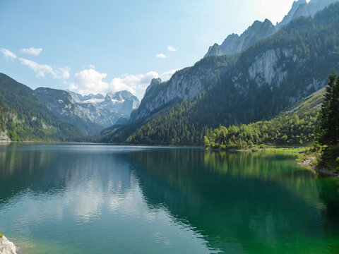 Panoramic view on Gosau lake, with Dachstein glacier in the back in Austrian Alps. The lake is surrounded by high mountains, overgrown with tall trees. Sun reflects on the surface. Serenity and calm © Chris