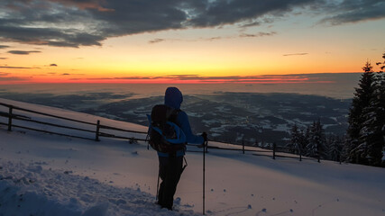 A man with hiking backpack and sticks standing at the snow covered peak of Schoeckl in Austrian Alps and admiring a sun rising above the horizon. The sky is bursting orange.Winter wonderland Day break