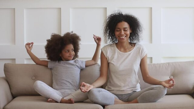African mom teacFunny african family young healthy mom teaching cute little kid daughter meditate together sit on sofa at home, calm mindful black mother and child girl do yoga exercise relax together