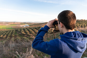 Unrecognized young boy standing in the mountain looking the landscape using binoculars.