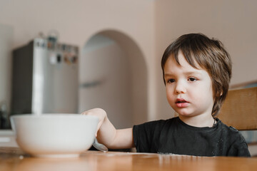 A cute little boy holding a spoon and having having breakfast at home