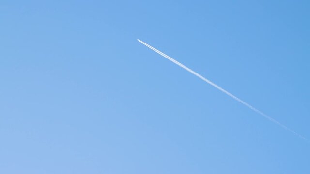 An airplane flies high above the ground leaving an even white trail. Blue sky, natural landscape illuminated by the sun. Beautiful background with clear weather and copy space. Time-lapse 4K video.