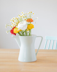 Bouquet of fresh ranunculus flowers and daisies in a mint green coffee pot from the 60ies and a chair in the same color and design on a wooden table.