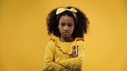 curly african american girl with crossed arms holding smartphone isolated on yellow