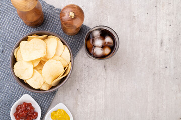 Potato chips in a bowl, pepper, salt, mustard, ketchup and a glass of soda. Light grey background with copy space