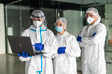 Medical workers in hazmat suit looking laptop inside clinic. Doctors and nurse with protective face masks talking about contagion peak inside hospital room.