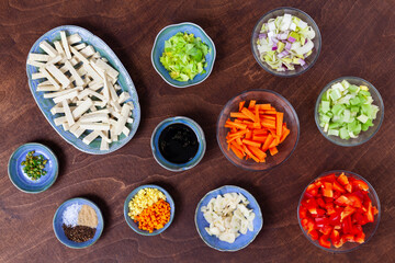 Flat lay of ready to make stir-fry ingredients: firm tofu, cut vegetables, soy sauce, spices in bowls.