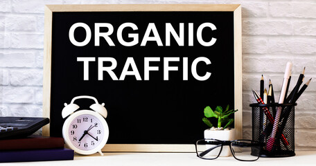 The words ORGANIC TRAFFIC is written on the chalkboard next to the white alarm clock, glasses, potted plant, and pencils in a stand.