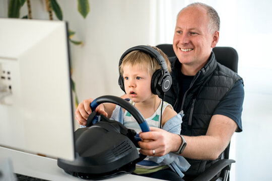 Small boy carefully watching his dad playing racing car game on the computer using a steering wheel
