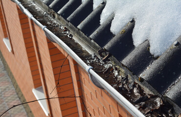 A close-up of a clogged, uncleaned roof gutter with last year fallen leaves and dirt while snow is...