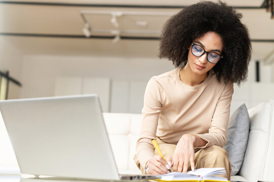 Smart young African American student or businesswoman sitting on the couch with laptop, studying from home, watching online webinar, taking notes to the notebook, making to do list, planning her week