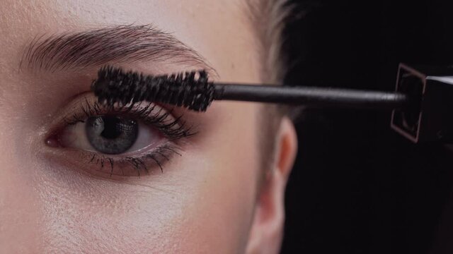 The makeup artist applies mascara to the model's eyelashes with a brush. Extreme close-up of the left eye on a black background.
