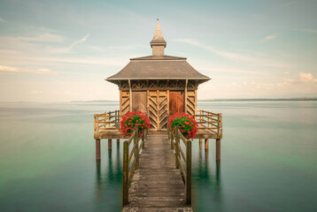 A small public bathhouse on Lake Geneva adorned with red flowers in the sunlight