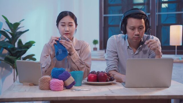 Business male freelance using laptop working at home office while female doing hobby by crochet handcraft in living room at night. Young Asian family Couple spend time together during lockdown