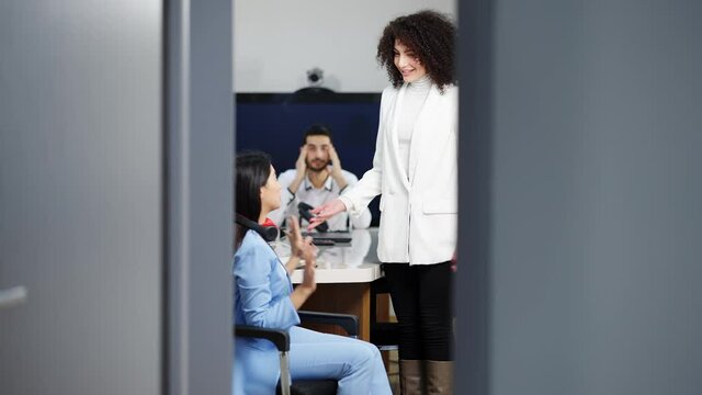 Wide shot of irritated young Caucasian woman approaching to Asian colleague listening music in headphones and banging fist on table. Shooting through doorway of multiethnic coworkers conflict in