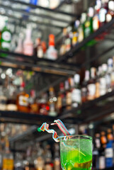 Green cocktail on the bar. Shelves with bottles of alcohol blurred in the background. Sharpness on a fruit cocktail with lemon.