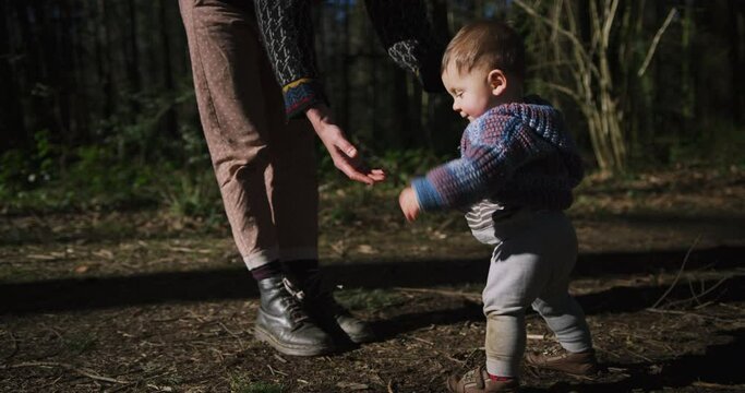 A young mother is helping her baby stand and walk in the woods on a sunny day of spring