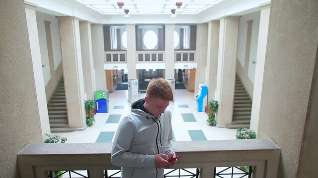 The red-haired guy is using the phone on the balcony of the dorm lobby, holding a gadget, typing a message. The use of mobile Internet in communication.