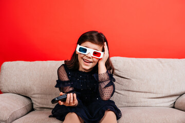 Happy child enjoying tv on sofa. Indoor shot of little girl in 3d glasses sitting on red background.