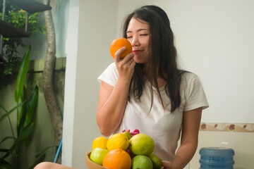 Fototapeta premium lifestyle portrait of young happy and cheerful girl holding orange smiling while sitting on kitchen board with fruit bowl in healthy nutrition concept