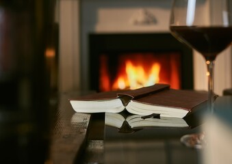 Opened book and red wine on table with fireplace in the background.