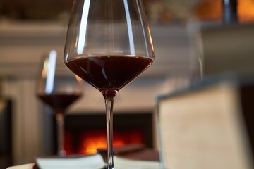 Two glasses of red wine with book and candle on table at home, fireplace in the background. Warm, dark colors.