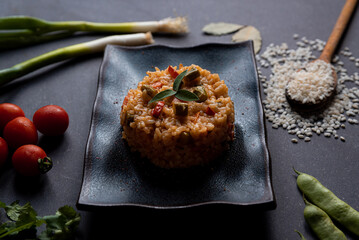 italian vegan spring risotto on black dish with asparagus on dark background. healthy and fresh springtime vegetables like carrots, cherry tomatoes and spring onions on black background