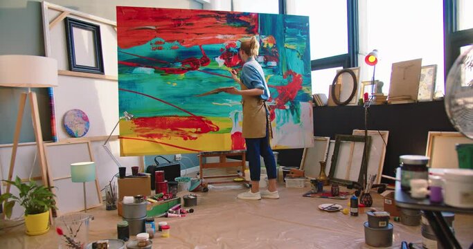 Rear of Caucasian young professional talented female painter working drawing on big canvas in workshop. Modern creating painting on large canva in art studio. Artist in apron working in gallery
