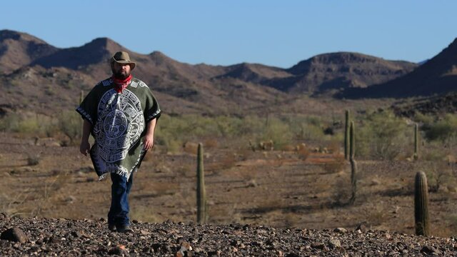 In the Sonoran desert, a desperado man wearing a poncho, red bandanna and a cowboy hat walks through a rugged landscape filled with rocks and saguaro cacti. In the distance, brown mountains are seen.