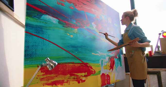Side view of Caucasian young pretty talented female painter working in art studio drawing on big canvas. Modern creating painting on large canva. Woman professional artist in apron working in workshop