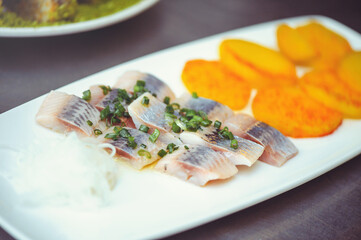 Delicious pieces of Icelandic herring with onions on a white plate. Raw salted herring on wooden background. Marinated herring fillet with grilled potatoes