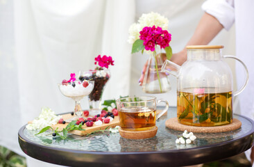 Hand with vase of blossom, glass teapot and cup of herbal tea, bowls with black current and sugar, mint leaves, raspberries and pink flowers.