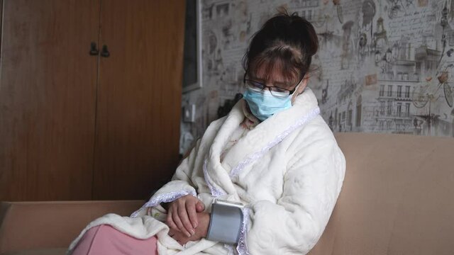 Mature woman in medical mask measuring pressure with digital tonometer, while sitting on sofa. Senior woman is taking care for health with hearth beat monitor. Healthcare and medical concept.