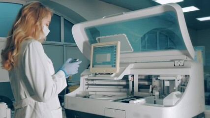 Woman in laboratory using. the mobile device receives information from the biochemical blood analyzer. Laboratory worker in a medical mask looks at a mobile phone