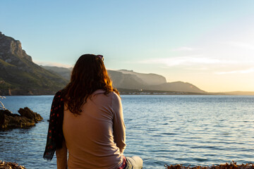A young and beautiful girl dressed in casual clothes looking at the sunset at a beautiful beach in Artà Mallorca Balearic Islands Spain with a impressive view of the sea and the Tramuntana mountains