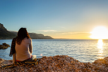 A young and beautiful girl dressed in casual clothes looking at the sunset at a beautiful beach in Artà Mallorca Balearic Islands Spain with a impressive view of the sea and the Tramuntana mountains
